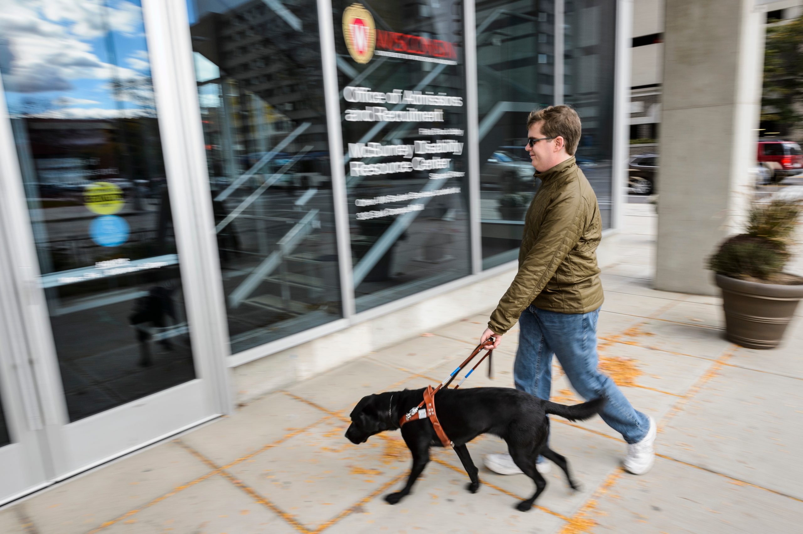Man in green jacket and blue jeans walking with his guide dog towards glass entrance doors of a building.